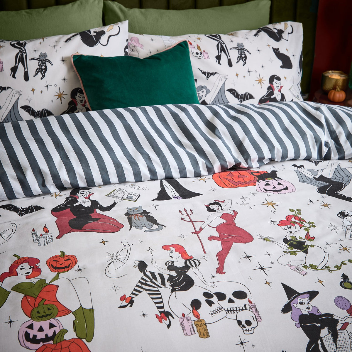 Reversible duvet cover on a bed, one side printed with Halloween pin-up characters, pumpkins and skulls on cream, the other black and white striped, styled with a green velvet cushion.