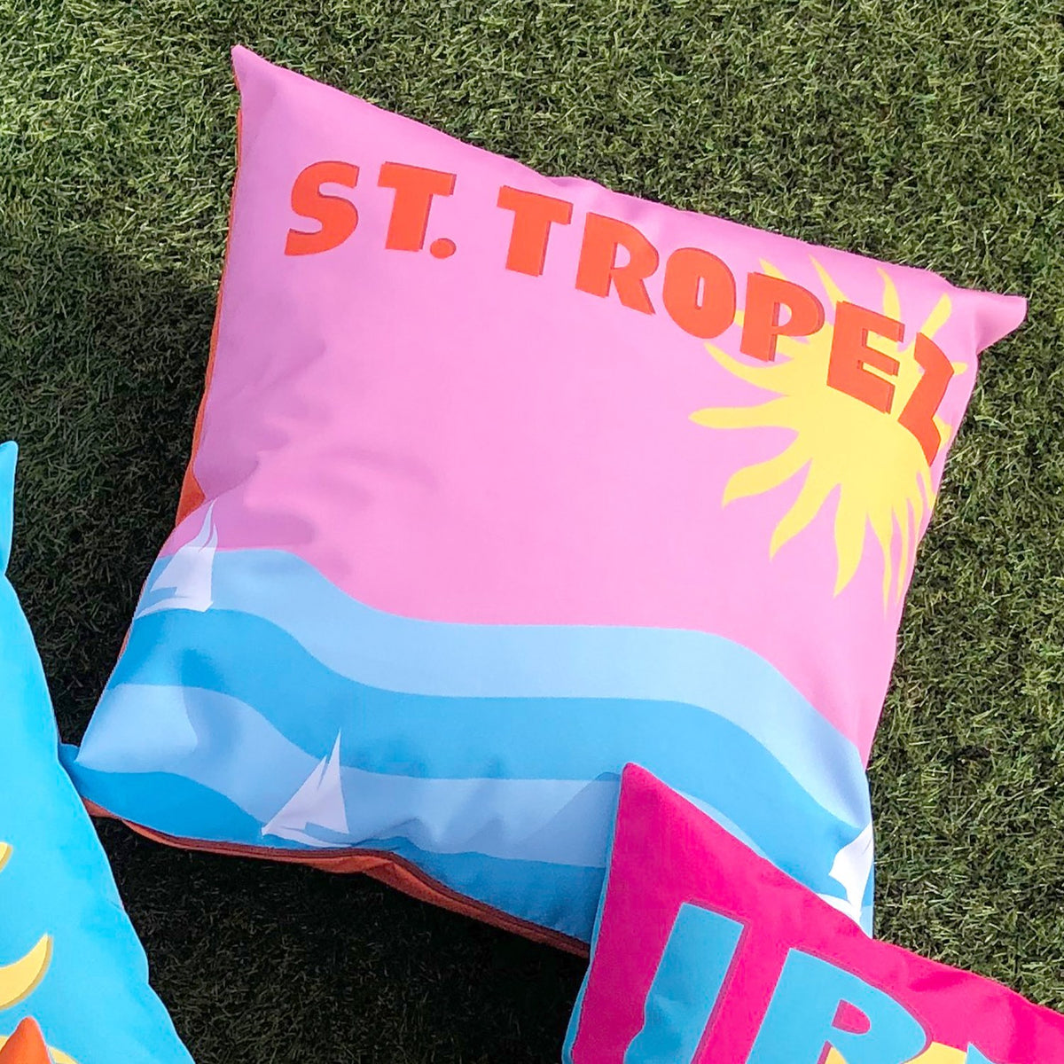 Square outdoor polyester cushion with pink background, orange "ST. TROPEZ" lettering, a sun graphic, and blue wave design with sailboats, shown on artificial grass.