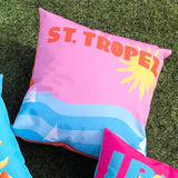 Square outdoor polyester cushion with pink background, orange "ST. TROPEZ" lettering, a sun graphic, and blue wave design with sailboats, shown on artificial grass.