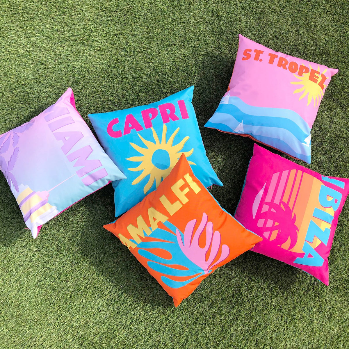 Square polyester outdoor cushion with pink sky, orange sun and blue wave design, "ST. TROPEZ" text, shown among other multicoloured city-print cushions on grass.