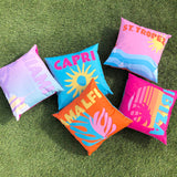 Square polyester outdoor cushion with pink sky, orange sun and blue wave design, "ST. TROPEZ" text, shown among other multicoloured city-print cushions on grass.