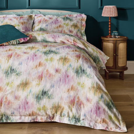 Trueno cotton duvet cover set with abstract watercolour brushstrokes in pink, purple, green and gold on cream, shown on a bed with matching pillowcases in a teal-walled bedroom.