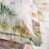 Close-up of the Trueno cotton duvet cover set showing an abstract watercolour print in pink, purple, green, gold and teal on a cream ground, with pillowcase edge and flat sheet visible.