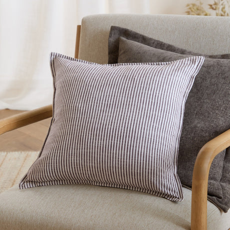 Truro Striped Cotton Cushion in chocolate brown and off-white pinstripe, square shape with piped edge, shown on a light upholstered armchair with grey cushions behind.