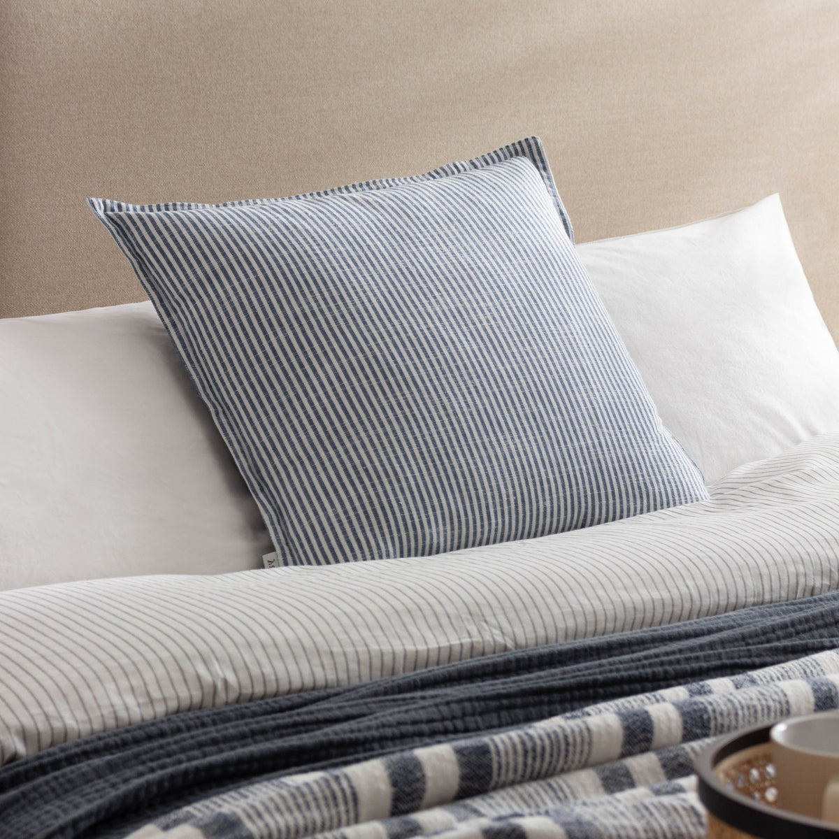 Truro Stripe Reversible Cotton Cushion in Skyline blue and white, shown on a bed layered with striped linen bedding and a woven throw.