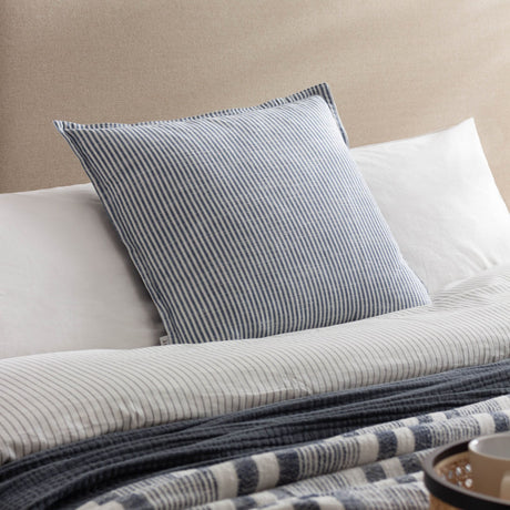 Truro Stripe Reversible Cotton Cushion in Skyline blue and white, shown on a bed layered with striped linen bedding and a woven throw.