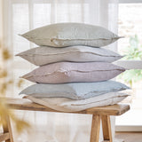 Truro Stripe Reversible Cotton Cushion in Skyline blue, stacked with striped cushions in varying tones on a wooden bench near a sheer-curtained window.