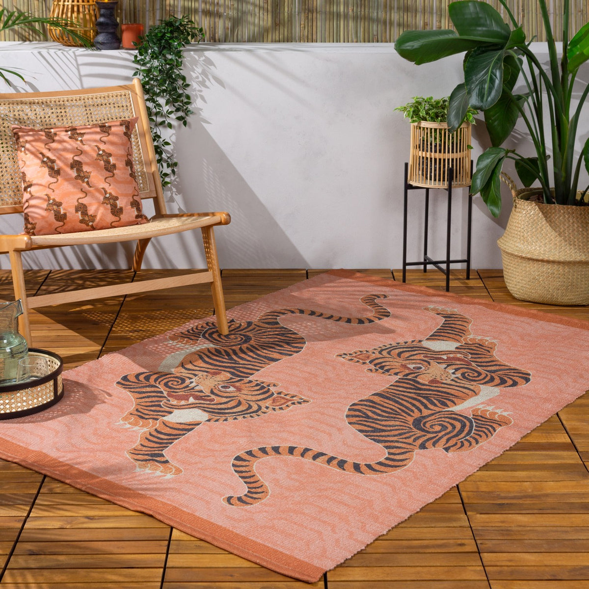 Tibetan Tiger Animal Polyester Rug in coral, flatweave design with two striped tigers, styled on a wooden deck with rattan chair and potted plants.