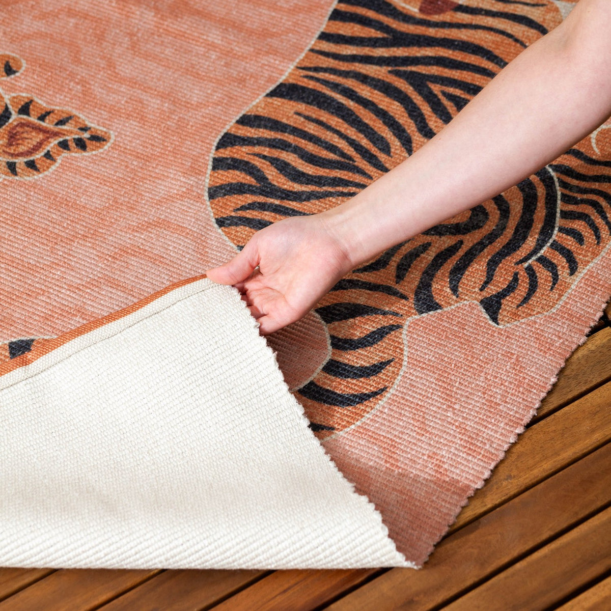 Hand folding back the corner of a coral Tibetan Tiger polyester flatweave rug, revealing its cream backing over a wooden deck.