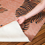 Hand folding back the corner of a coral Tibetan Tiger polyester flatweave rug, revealing its cream backing over a wooden deck.