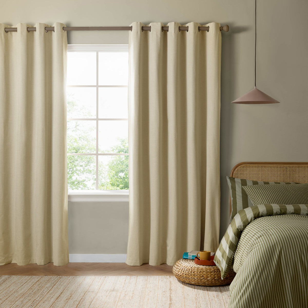 Custard cream eyelet blackout curtains in woven fabric, hung on a wood pole either side of a window in a bedroom with green striped bedding.