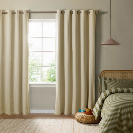 Custard cream eyelet blackout curtains in woven fabric, hung on a wood pole either side of a window in a bedroom with green striped bedding.