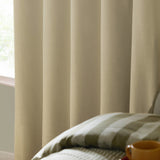 Custard cream eyelet blackout curtains in woven fabric, shown draped beside a bedroom window with a striped green and cream bedspread nearby.