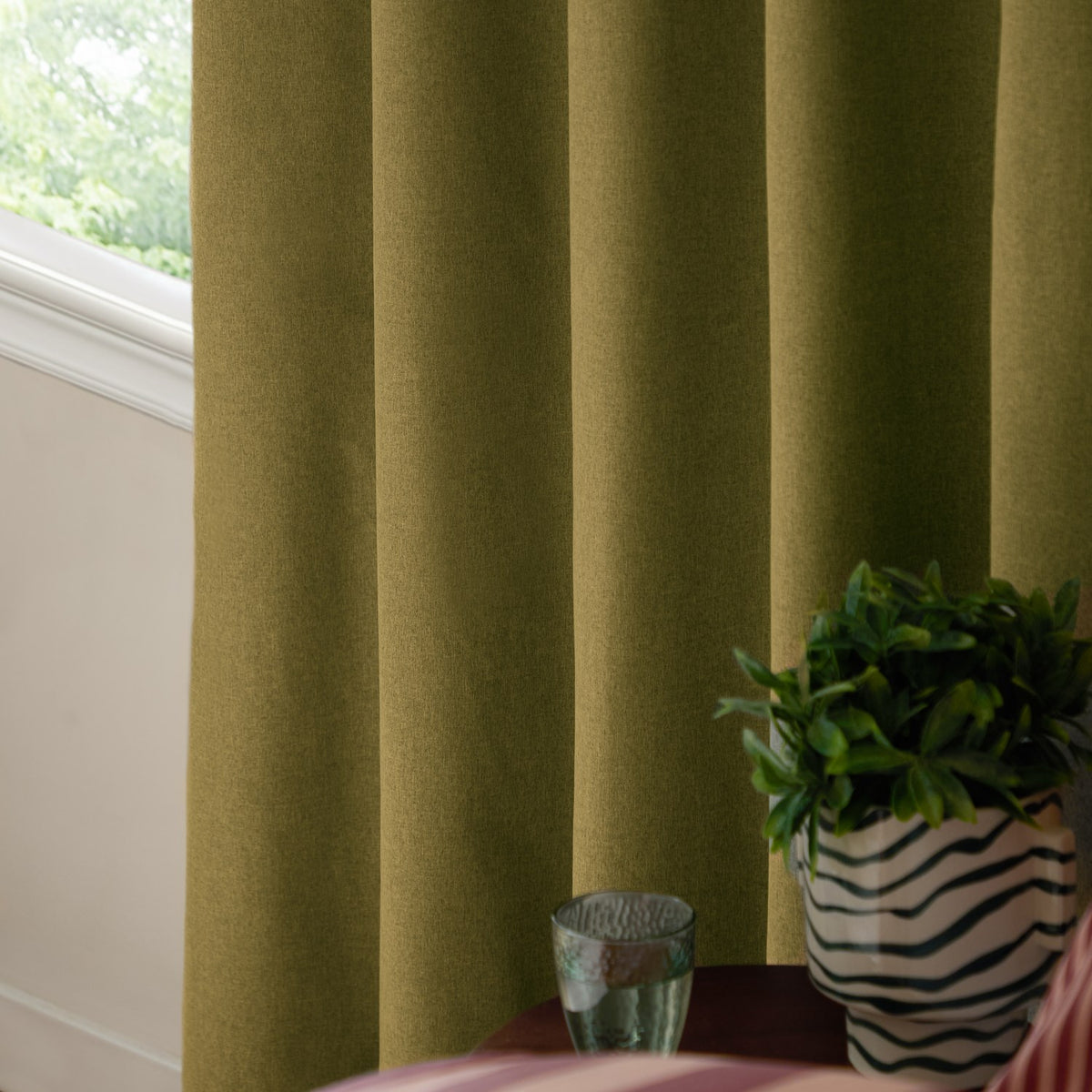 Forest green woven eyelet blackout curtains hanging in soft folds beside a window, styled with a side table, glass tumbler, and striped plant pot.