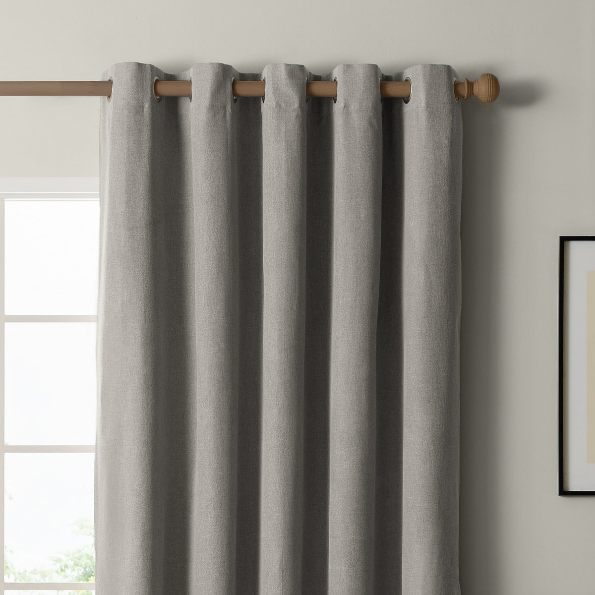 Grey eyelet blackout curtain in textured woven polyester, hung on a wooden pole with metal grommets, shown against a light wall beside a window.