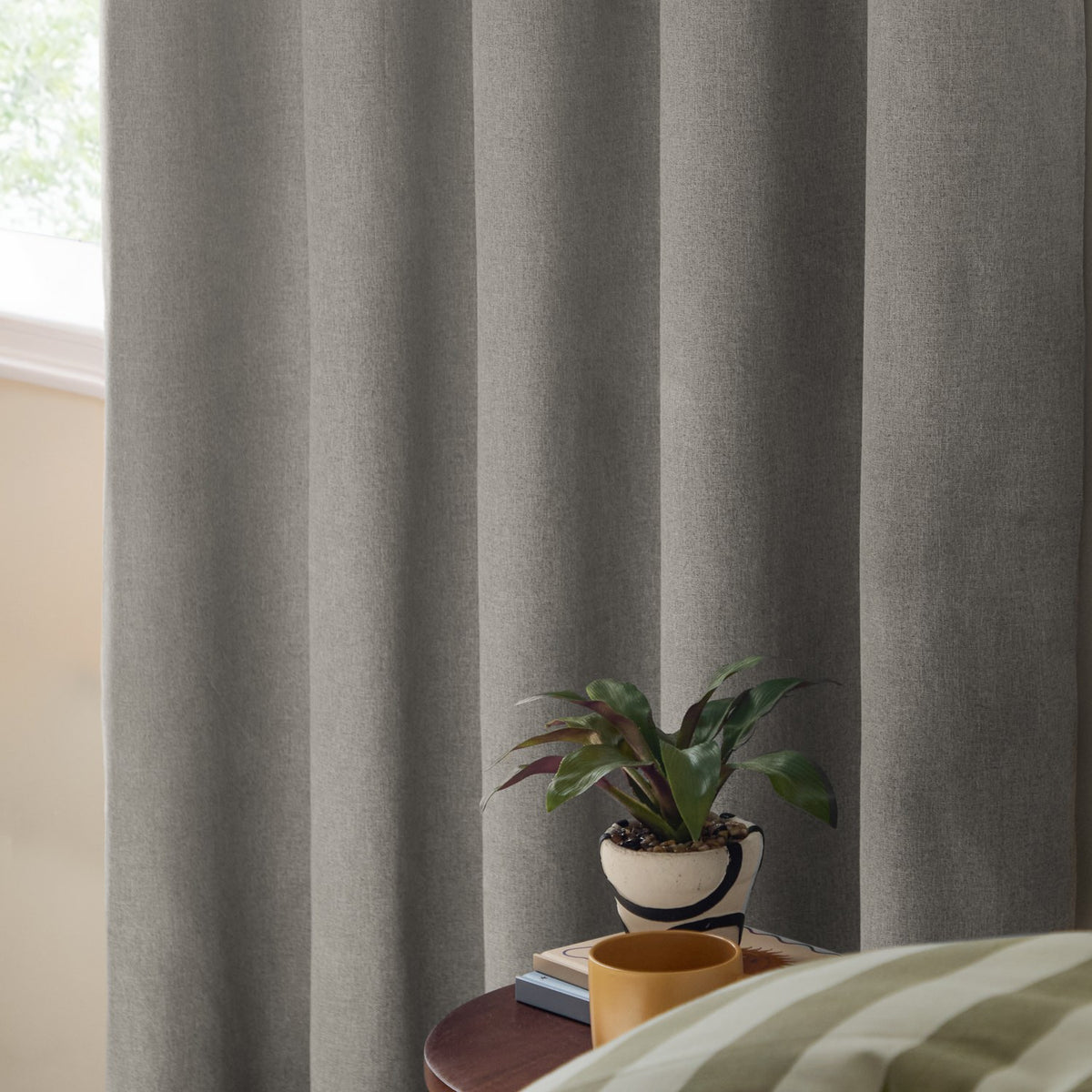 Grey woven eyelet blackout curtains hanging in draped folds beside a window, shown in a bedroom setting with a side table, plant, and mug.