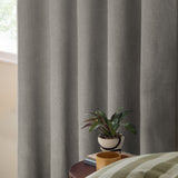 Grey woven eyelet blackout curtains hanging in draped folds beside a window, shown in a bedroom setting with a side table, plant, and mug.