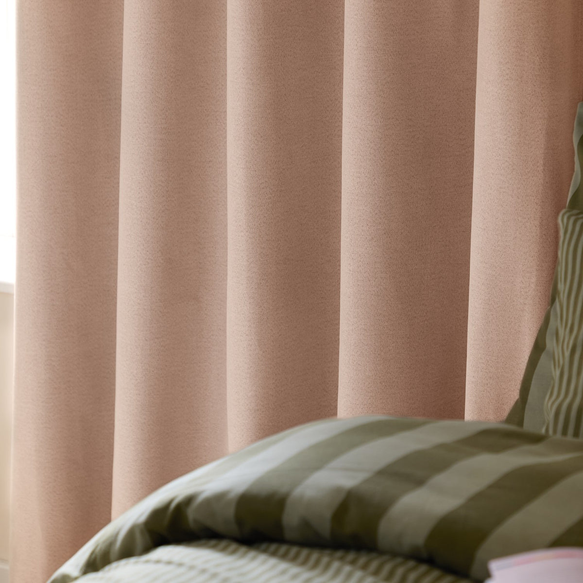 Blush pink woven eyelet blackout curtains hanging in soft folds beside a bed with olive green striped bedding.