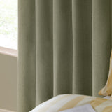 The Ultimate Blackout Plain Woven Eyelet Curtains in sage green, shown in soft folds by a window, with a striped bed cushion visible alongside.