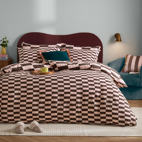 The Ultimate Checkerboard Check Duvet Cover Set in Berry Brownie on a bed with matching pillowcases, pink and brown check pattern, burgundy headboard, and blue armchair in a bedroom setting.