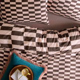 The Ultimate Checkerboard Duvet Cover Set in Berry Brownie, styled on a bed with pink and brown check pillowcases, striped edge trim, and a teal fringed cushion with a breakfast tray.