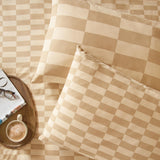 Checkerboard cotton blend pillowcases in caramel and cream on matching bedding, with a wooden tray, coffee cup, glasses, and open book alongside.
