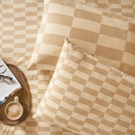 Checkerboard cotton blend pillowcases in caramel and cream on matching bedding, with a wooden tray, coffee cup, glasses, and open book alongside.