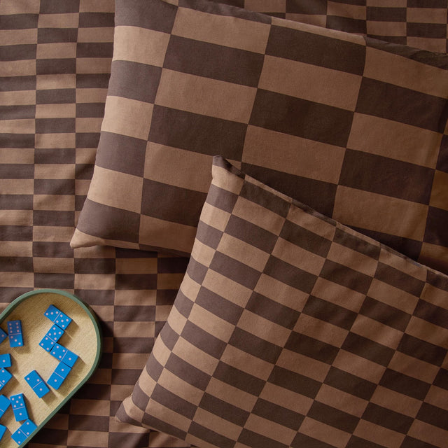 Ultimate Checkerboard cotton blend pillowcase pair in Chocolate Truffle, reversible oblong checkerboard design in two brown tones, styled on bedding with dominoes alongside.