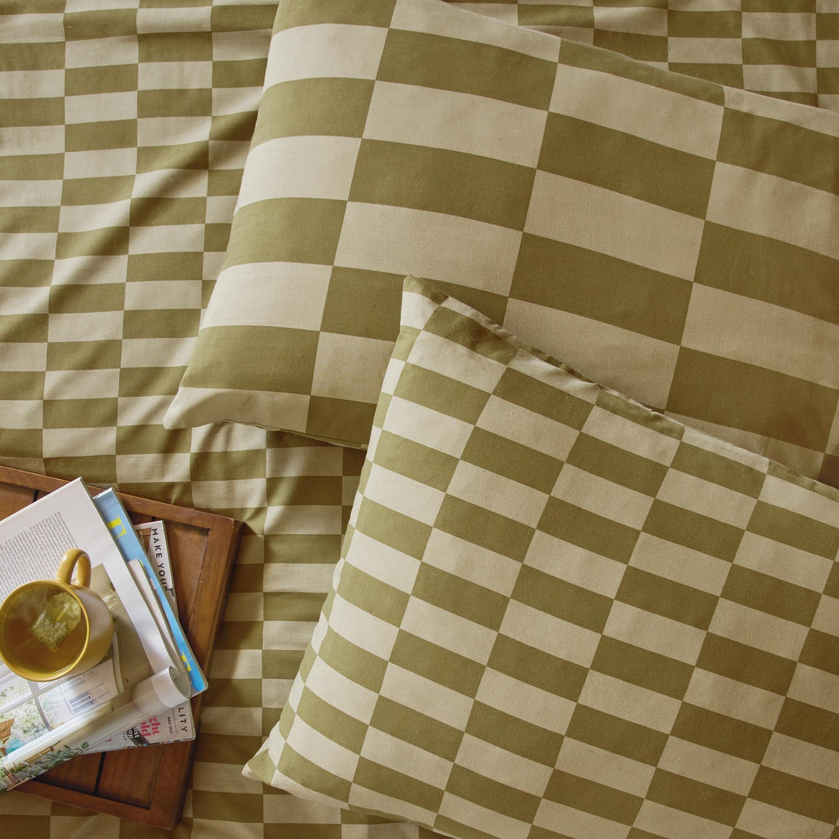 Green and cream checkerboard cotton blend pillowcase pair in Green Tea, shown on a bed with matching duvet, styled with a tray, mug, and magazines.