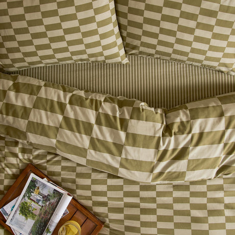 Green Tea checkerboard cotton blend pillowcase pair on a bedspread with matching striped and block-check patterns, styled with a bedside tray and magazine.