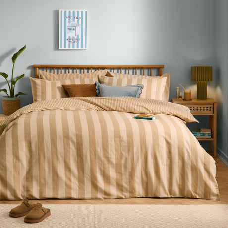 The Ultimate Stripe Reversible Duvet Cover Set in Caramel Latte dresses a bed with beige and cream striped bedding, styled with cushions in a light blue bedroom.
