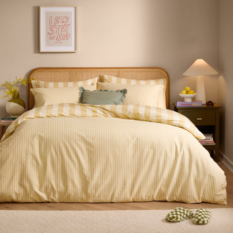 The Ultimate Stripe Reversible Duvet Cover Set in Custard Cream dresses a bed with striped pillowcases and a matching cover in a rattan-headboard bedroom.