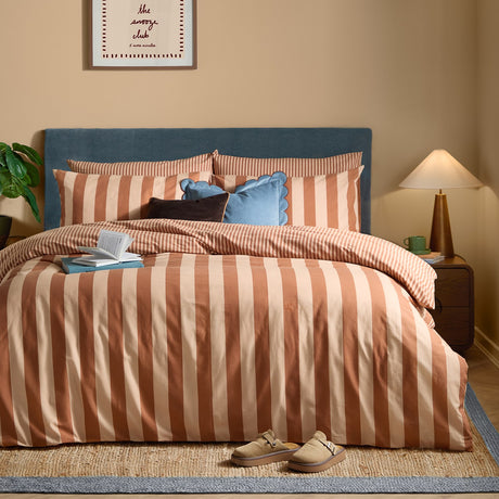 The Ultimate Stripe Reversible Duvet Cover Set in Pumpkin Spice, wide rust and cream stripes dressed on a bed with matching pillowcases in a bedroom with a blue upholstered headboard.