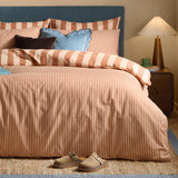 The Ultimate Stripe Reversible Duvet Cover Set in Pumpkin Spice dresses a bed with rust and cream stripes, styled with blue scalloped and velvet cushions in a bedroom setting.
