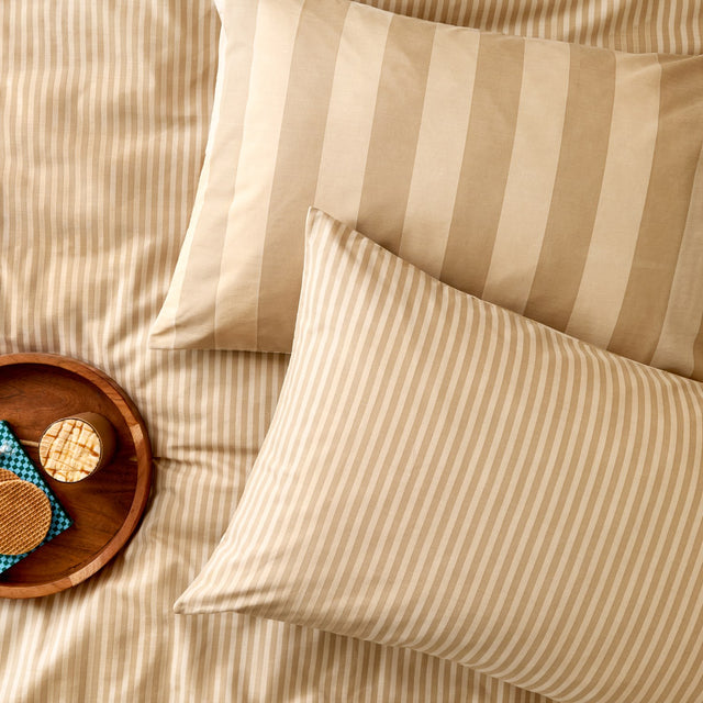 The Ultimate Stripe Cotton Pillowcase Pair in Caramel Latte, styled on a bed with narrow and bold striped sides, alongside a wooden tray of snacks.