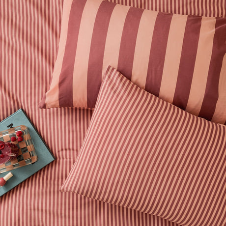 Cotton-blend reversible pillowcase pair in Raspberry Smoothie, styled on a matching striped bed with broad and fine pink and brick red stripes on each side.