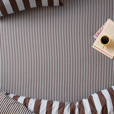 The Ultimate Stripe Cotton Blend Fitted Sheet in Blueberry Muffin, brown and white pinstripe fitted sheet on a bed with matching pillowcase, styled with a book and coffee cup.