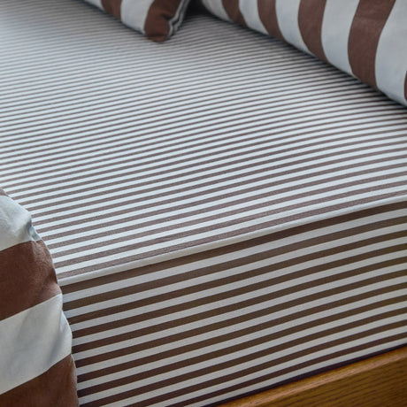 Close-up of The Ultimate Stripe Cotton Blend Fitted Sheet on a mattress, showing a narrow brown and white pinstripe pattern with matching striped pillowcases in the background.