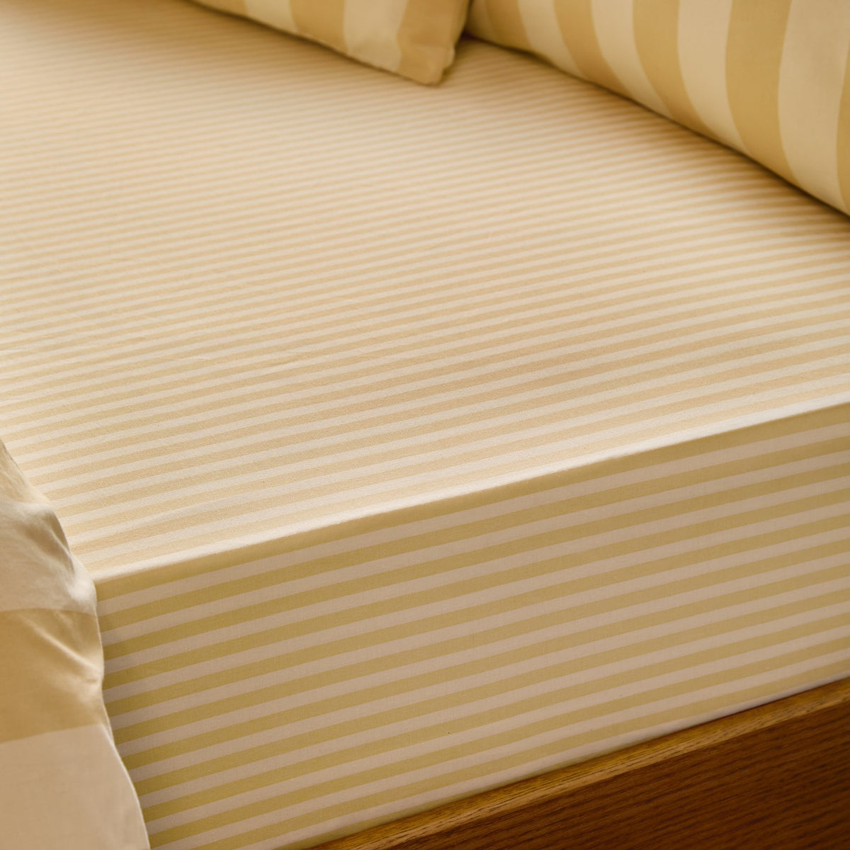 Fitted sheet in custard cream tonal stripe cotton-blend fabric, shown close-up on a mattress corner with matching striped pillowcases in a bedroom setting.