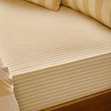 Fitted sheet in custard cream tonal stripe cotton-blend fabric, shown close-up on a mattress corner with matching striped pillowcases in a bedroom setting.
