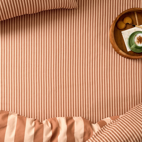 Pumpkin Spice Ultimate Stripe cotton blend fitted sheet on a bed with tonal orange-rust stripes, striped duvet fold, pillow, and a wooden tray of coffee and biscuits.