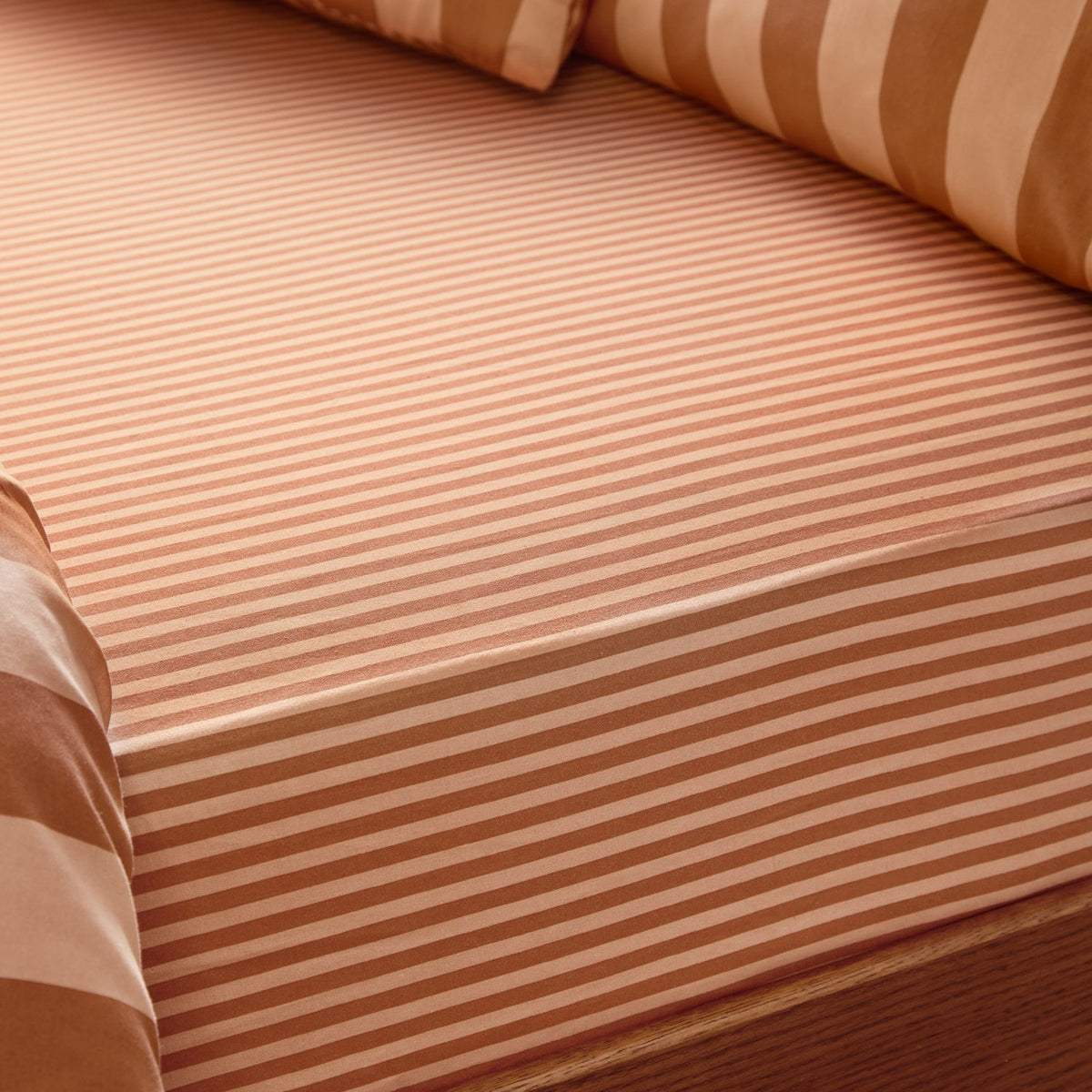 Close-up of The Ultimate Stripe Cotton Blend Fitted Sheet in Pumpkin Spice, showing fine tonal orange-rust stripes on a fitted corner, with a wider striped pillow behind on a bed.