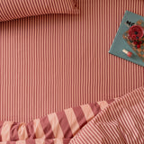 The Ultimate Stripe Cotton Blend Fitted Sheet in Raspberry Smoothie, styled on a bed with matching pinstriped pillowcase and duvet, alongside a book and drink.