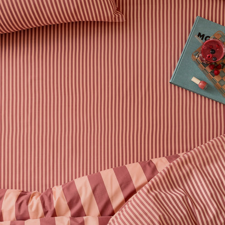 The Ultimate Stripe Cotton Blend Fitted Sheet in Raspberry Smoothie, styled on a bed with matching pinstriped pillowcase and duvet, alongside a book and drink.