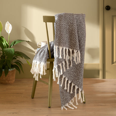 Tweedie Herringbone Polyester Throw in charcoal grey draped over a chair, showing the tweed-like chevron weave and twisted white fringe tassels.