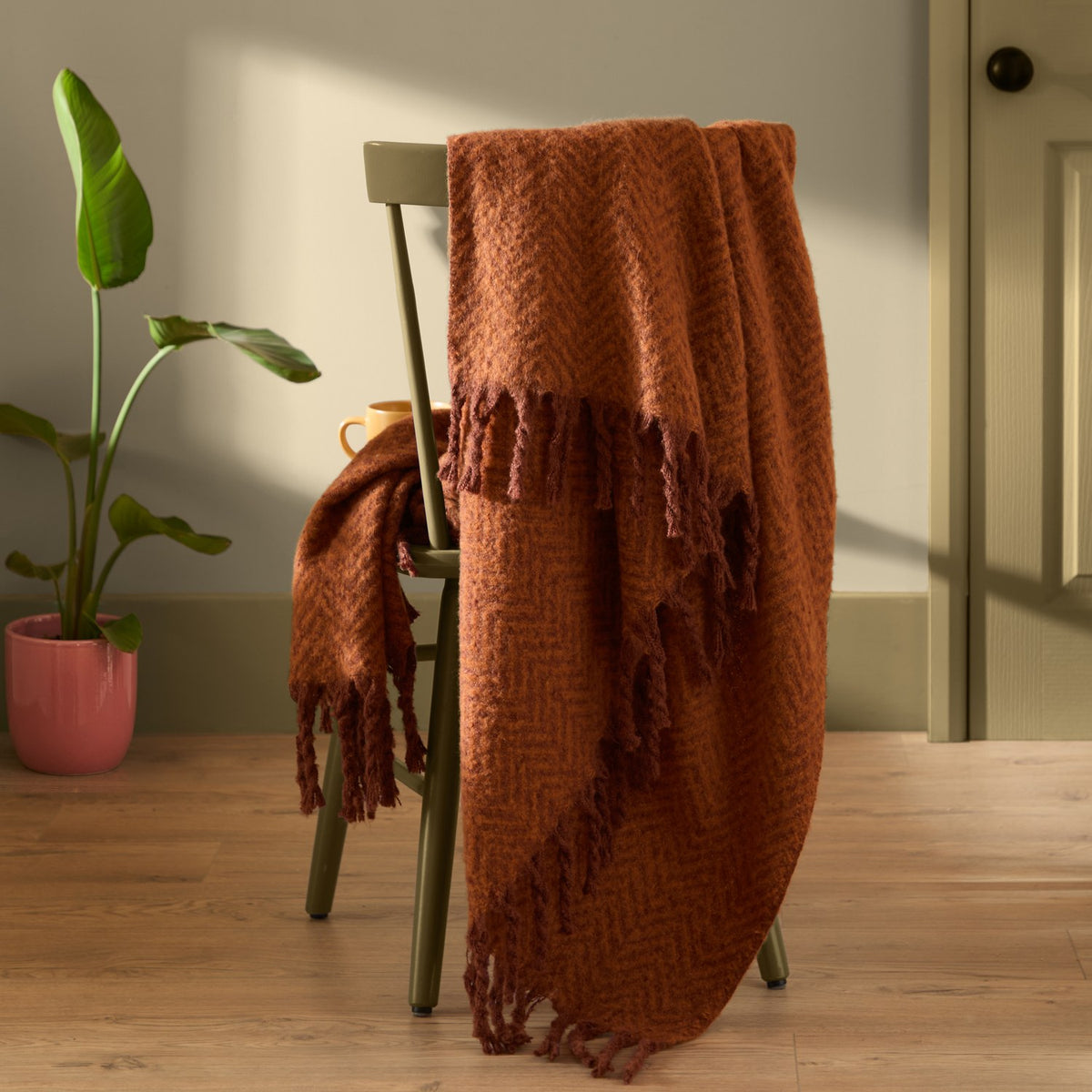 Tweedie Herringbone Polyester Throw in cinnamon rust draped over a green chair, showing its woven pattern and fringed edge in a living room setting.