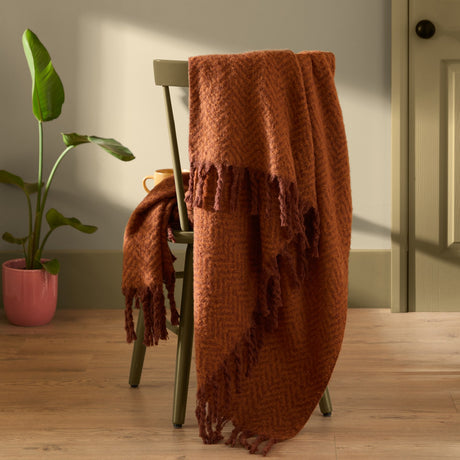 Tweedie Herringbone Polyester Throw in cinnamon rust draped over a green chair, showing its woven pattern and fringed edge in a living room setting.