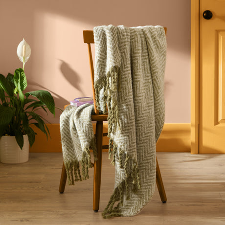 Tweedie Herringbone Polyester Throw in green and cream, draped over a wooden chair with fringed edges, styled in a living room with a plant and warm walls.