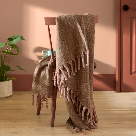 Tweedie Herringbone Polyester Throw in mocha brown draped over a chair, showing its chevron weave and long fringed edges in a warm-toned room.