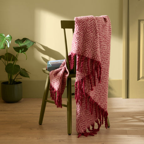 Tweedie Herringbone Polyester Throw in pink and burgundy with fringed ends, draped over a green wooden chair beside a potted plant.